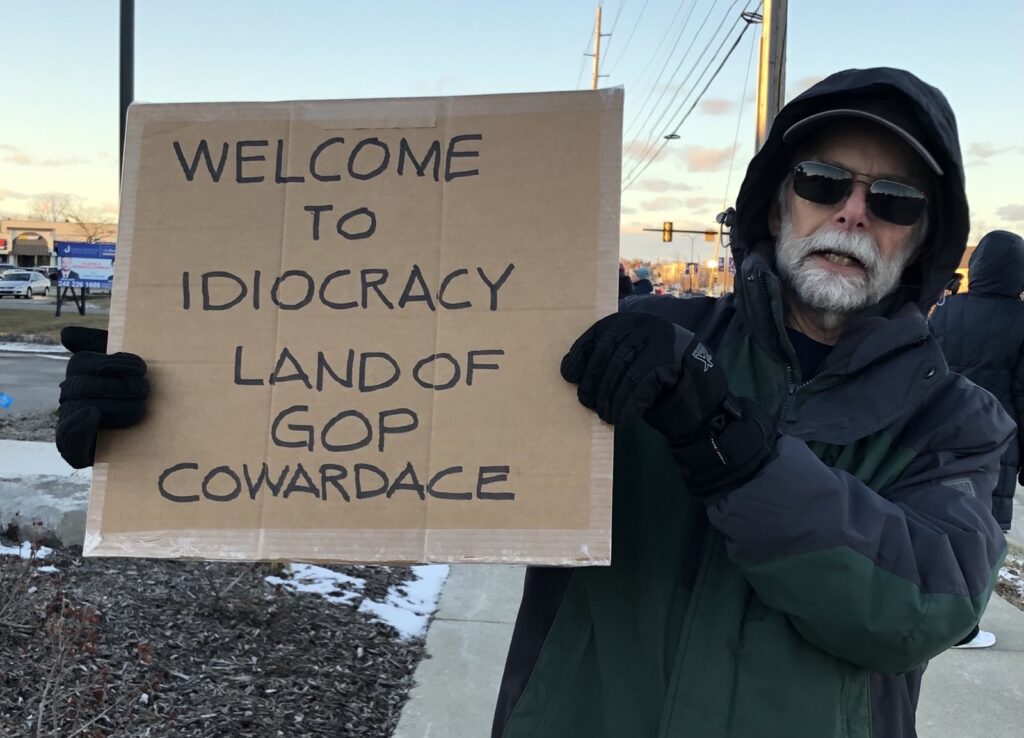 A man holding a cardboard sign that says "Welcome to Idocracy Land of GOP Cowardice"