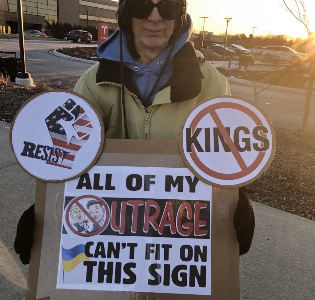 A woman holding a sign that says "All of my outrage can't fit on this sign" with another sign that says "No Kings" and another of a raised fist that says "Resist"