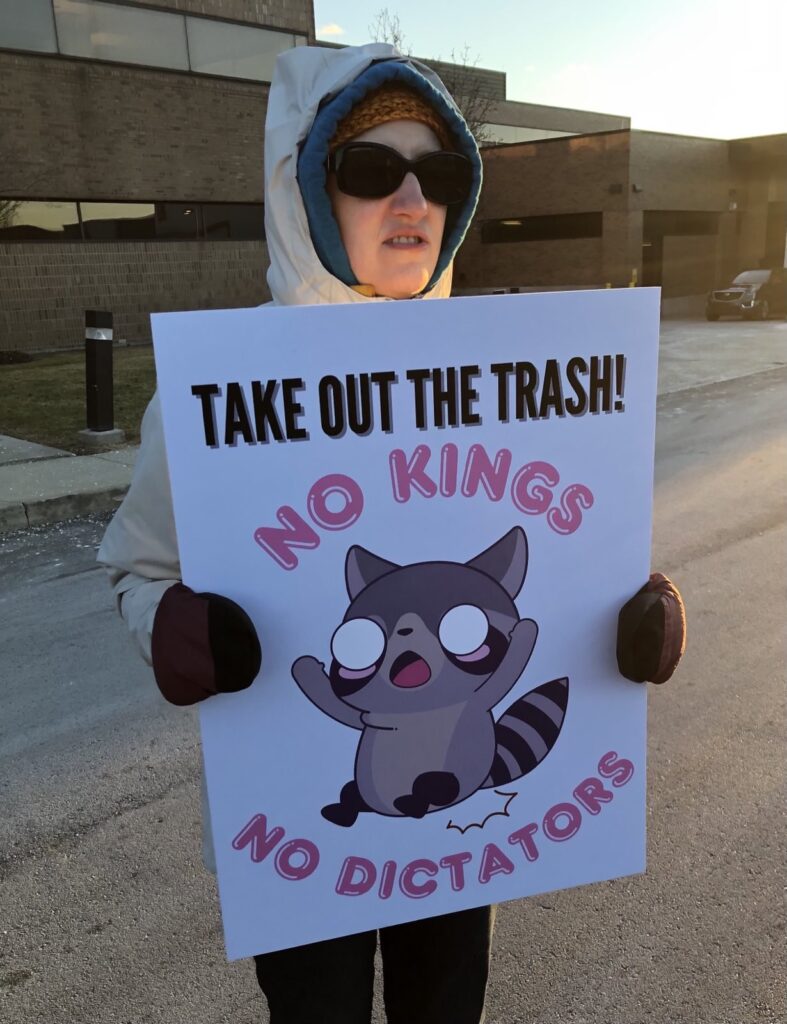 A woman holding a sign that says "Take out the trash!! No kings no dictators"